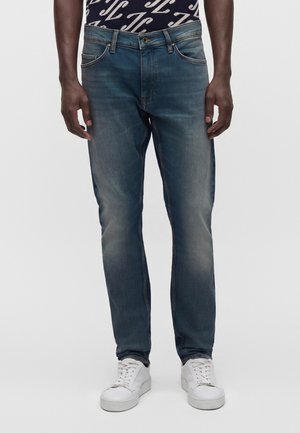 Mann trägt mittelblaue ausgewaschene Jeans und weiße Turnschuhe und steht vor einem schlichten hellen Hintergrund.