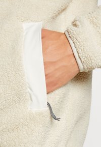 Tela de forro color crema con una textura suave; presenta un ribete de satén blanco en el bolsillo y un detalle de cordón con rayas en blanco y negro.
