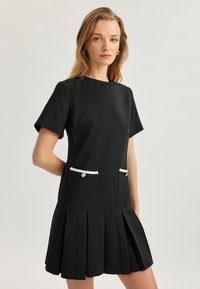 Vestido negro de manga corta con falda plisada, que presenta bolsillos con detalles en blanco y botones dorados. Tejido suave con una silueta limpia.