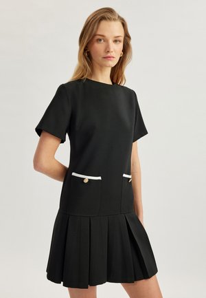 Schwarzes kurzes Kleid mit kurzen Ärmeln und gepleatem Rock, mit weiß abgesetzten Taschen und goldenen Knöpfen. Glattes Material mit klarer Silhouette.