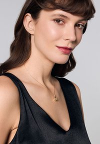 Złoty naszyjnik z zawieszką w kształcie łzy na delikatnym łańcuszku, noszony na głębokim dekolcie w kształcie litery V czarnej bluzki. Gładka tekstura, prosty design, minimalne akcenty.