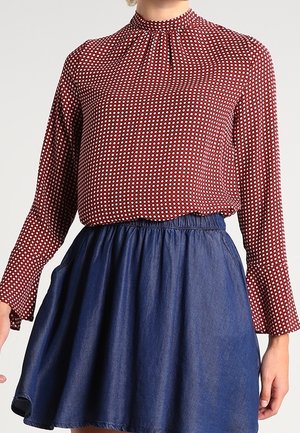 Vrouw draagt een rood, gebloemd, blouse met lange mouwen, ingestopt in een donkerblauwe, hooggetailleerde uitlopende rok.