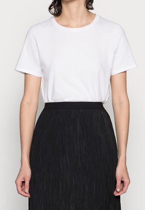 Femme portant un t-shirt blanc uni à manches courtes rentré dans une jupe noire texturée taille haute sur un fond neutre.