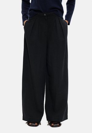 Osoba nosząca luźne, czarne spodnie o szerokich nogawkach z bocznymi kieszeniami, zestawione z czarnymi sandałami na otwartym palcu oraz granatową bluzką z długim rękawem.