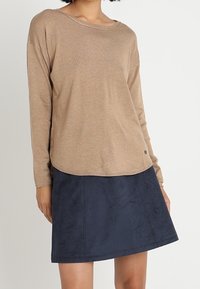 Pull beige à manches longues avec un large col et des boutons sur le côté, associé à une jupe trapèze en daim bleu marine avec des détails de coutures.