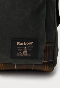 Dark green fabric with a black Barbour logo patch depicting a lighthouse, above a brown and orange plaid patterned fabric section.