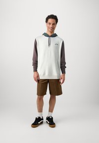 Light gray hoodie with dark brown sleeves, front pocket, and drawstring. Brown shorts and black sneakers with gum soles, stance casual.