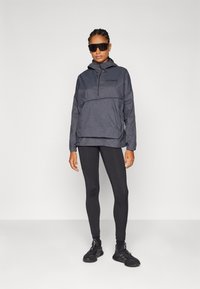 Pulover de culoare gri închis, cu fermoar pe un sfert și glugă, asortat cu colanți negri și pantofi sport negri, accentuat de ochelari de soare mari.