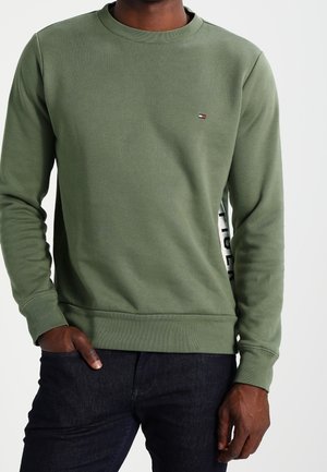 Mann trägt ein olivgrünes Langarm-Sweatshirt mit kleinem Logo auf der Brust und schwarze Jeans, steht mit einer Hand in der Tasche.