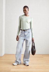Lys grønn langermet topp, høy midje lyseblå jeans, svart belte, svart veske og grå joggesko på et tregulv mot en lys vegg.