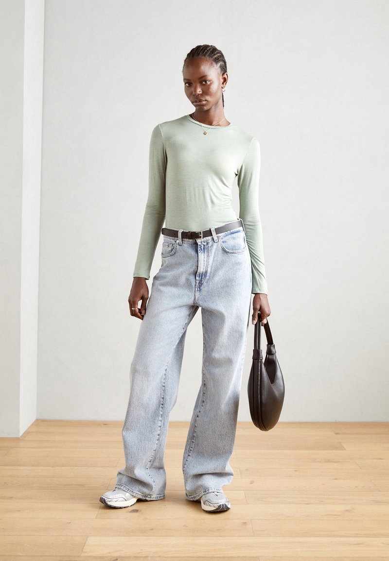 Lys grønn langermet topp, høy midje lyseblå jeans, svart belte, svart veske og grå joggesko på et tregulv mot en lys vegg.