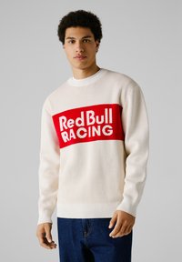 Cremfarbener Pullover mit einem auffälligen roten horizontalen Streifen, der "Red Bull RACING" in Weiß zeigt; gerippter Kragen und Bündchen; weiche Stricktextur.