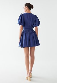 Mornarsko moder kratek obleke s pufastimi rokavi, prilegajočim pasom, nabranim krilom in robom z oglatimi detajli. Narejena iz lahkega materiala.