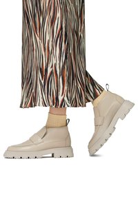 Botas de tobillo de cuero beige con una correa, suela texturizada y gruesa, y una lengüeta. Combinadas con una falda con estampado y calcetines amarillos claros.