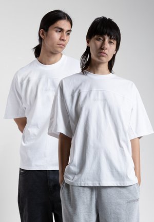 Two individuals wearing white oversized t-shirts with a subtle design on the chest, set against a neutral background.