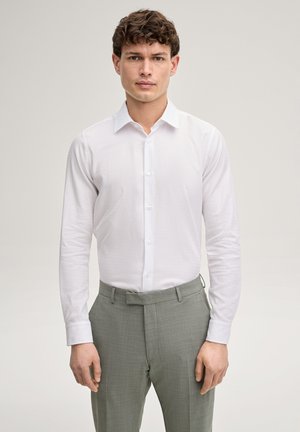 Junger Mann, der ein eng anliegendes weißes Hemd und hellgraue Hose trägt, vor einem schlichten hellen Hintergrund stehend.