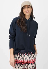 Felpa blu navy con cappuccio e cordoncino con punte argentate, abbinata a pantaloni con motivi geometrici in rosso, nero e beige.