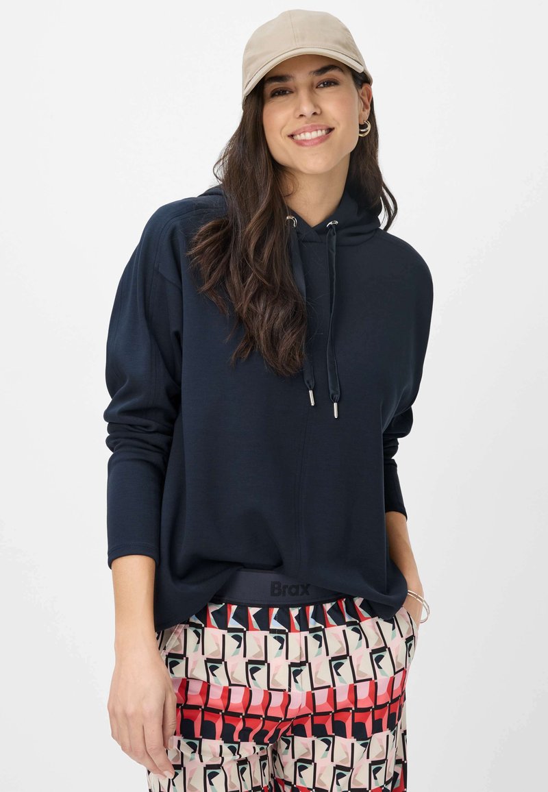 Felpa blu navy con cappuccio e cordoncino con punte argentate, abbinata a pantaloni con motivi geometrici in rosso, nero e beige.