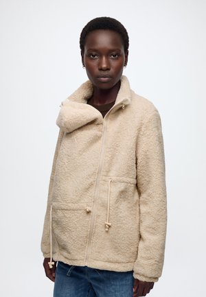Femme portant une veste polaire beige zippée avec des cordons et un col montant, associée à un jean bleu, debout devant un fond blanc uni.