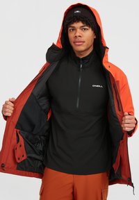 Sort zip-up mid-layer trøje, der er lagdelt under en orange hættejakke, med en glat tekstur og logo-detalje på brystet.