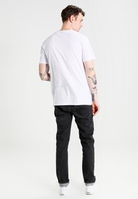 Hvid t-shirt med korte ærmer iført mørke jeans og hvide sneakers. Synlige tatoveringer på armene, afslappet pasform, ensfarvet design, ingen mønstre.
