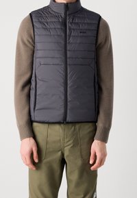 Grå pufferväst med hög krage, dragkedja framtill och vertikala sömmar; matchad med en beige tröja och gröna byxor. Tunn textur, lätt design.