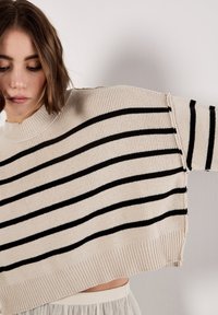 Pull rayé à coupe décontractée, tricot texturé en rayures beige et noires, col rond large, et design court avec des poignets côtelés.