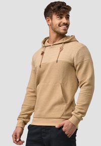 Sudadera beige de canalé con un bolsillo frontal, cordones y acentos metálicos. Presenta un corte holgado y una textura suave. Combinada con pantalones oscuros.
