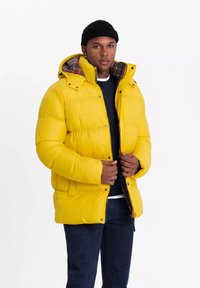 Gul pufferjakke med hætte og et ternet for. Designet har en lynlås, trykknapper og en polstret tekstur. Bæres over en mørk sweater.