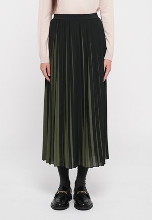 Mujer que lleva una falda midi plisada negra y verde oliva, camiseta beige de manga larga, calcetines oscuros y mocasines negros con detalle de cadena dorada.
