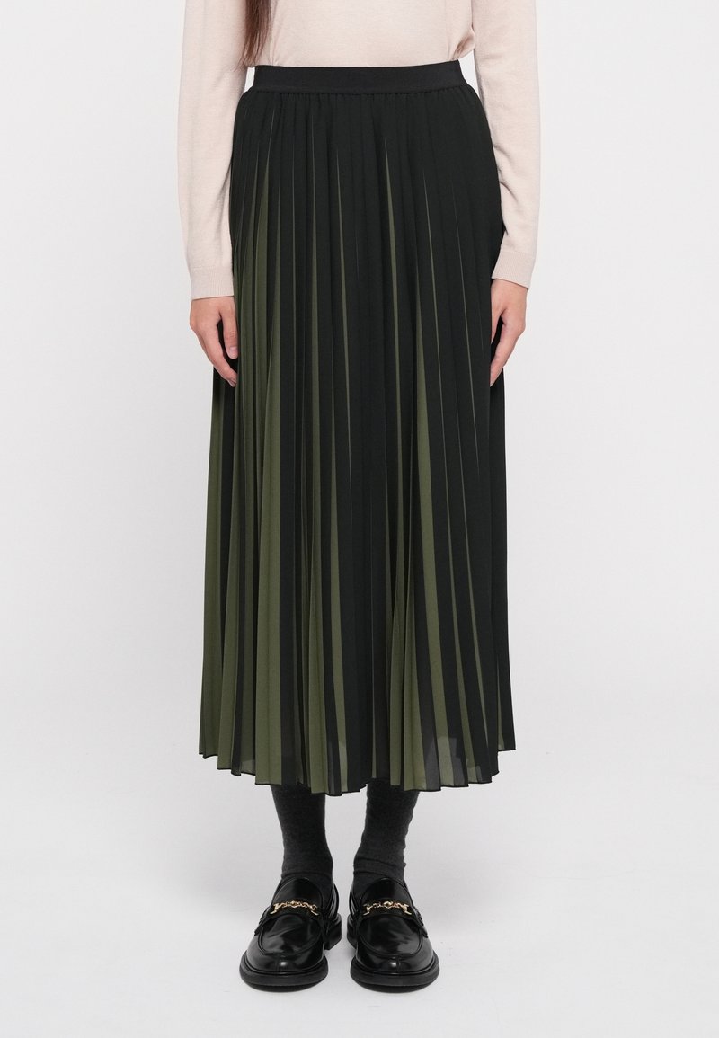 Femme portant une jupe midi plissée noire et vert olive, un haut beige à manches longues, des chaussettes foncées et des mocassins noirs avec détail de chaîne dorée.