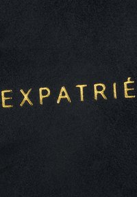 Złote haftowane litery tworzące słowo „EXPATRIÉ” na czarnym tle z tkaniny.