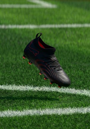 Fekete és piros focicipő a levegőben, egy zöld fűvel borított, fehér vonalakkal jelölt pálya felett.