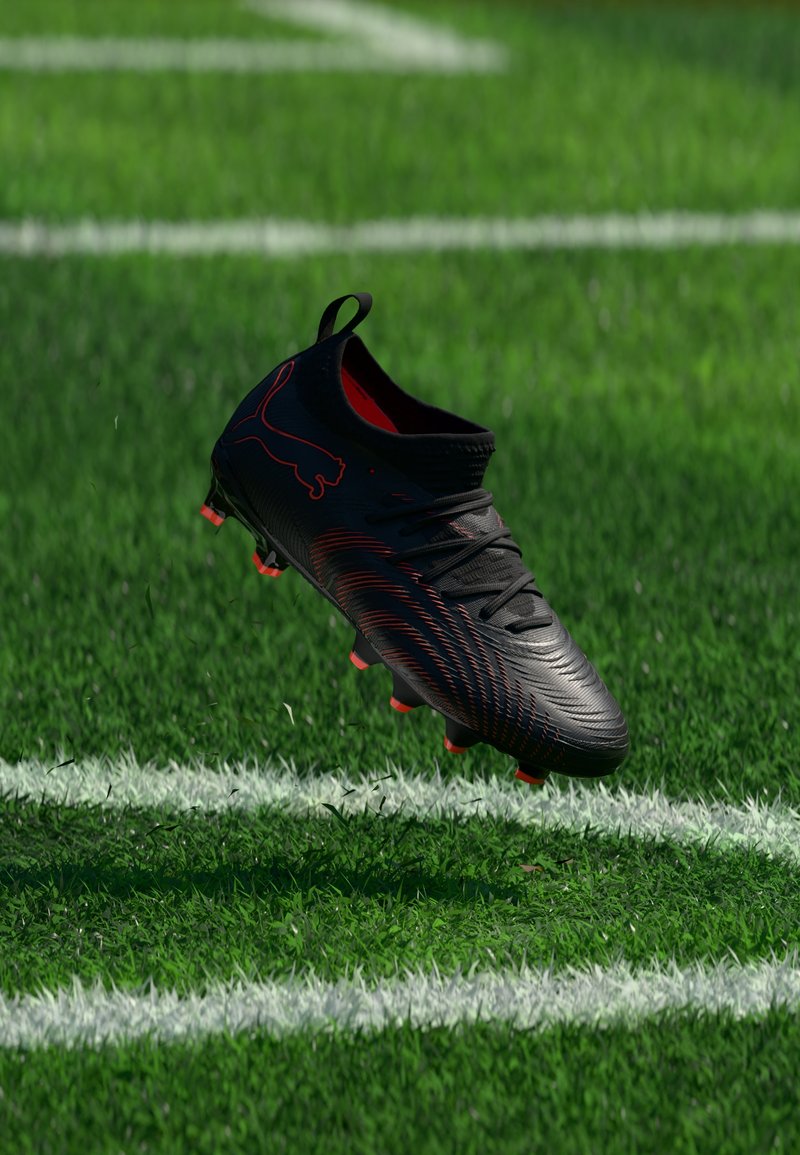 Chaussure de football noire et rouge en l'air au-dessus d'un terrain herbeux vert marqué de lignes blanches.