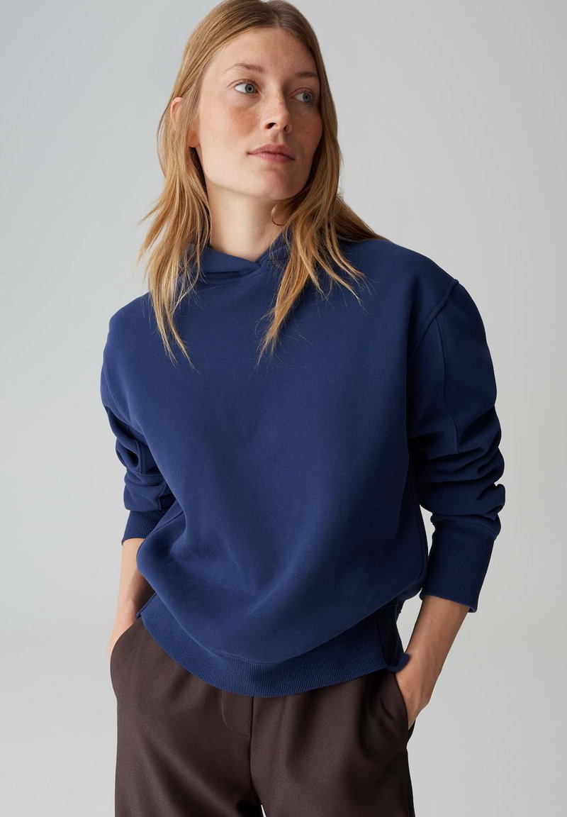 Marineblauer Hoodie mit lockerem Schnitt, gerippten Bündchen und Kängurutasche. Kombiniert mit weit geschnittenen braunen Hosen. Glatte Textur.