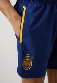 Calções azuis marinhos com um detalhe amarelo, bolso com fecho éclair e o emblema da Espanha apresentando uma coroa e uma estrela, feitos de um material leve e suave.