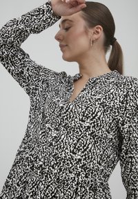Schwarz-weiß gemustertes Kleid mit Kragen, langen Ärmeln, Knopfleiste und gerafftem Kleidungsbund. Glattes Material mit strukturiertem Druckdesign.