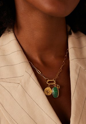 Gold chain necklace with compass and green stone pendants worn on a person in a beige pinstripe collar garment.