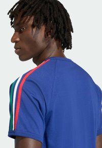 Muž s krátkymi dredmi, oblečený v modrom tričku so zelenými, bielymi a červenými pásmi na jednom ramene, otočený doľava na bielom pozadí.