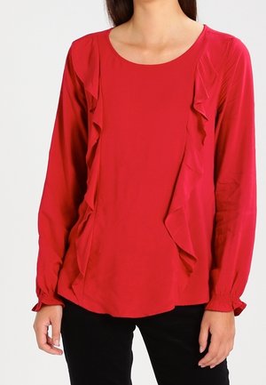 Nainen pukeutunut pitkähihainen punainen pusero, jossa on röyhelöyksityiskohtia edessä ja kerätyt kalvosimet, yhdistettynä mustiin housuihin.