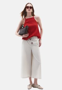 Top in maglia rossa con spalline sottili, pantaloni beige a gamba larga, borsa a tracolla nera intrecciata e scarpe slip-on color sabbia chiara. Occhiali da sole marroni.