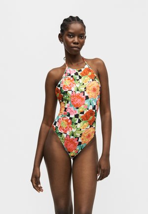 Femeie în picioare purtând un costum de baie întreg cu decolteu halter și un model floral mare, colorat, pe un fundal în carouri.