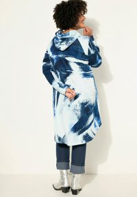 Manteau long à capuche dans un motif abstrait bleu et blanc, offrant une coupe décontractée et une fente latérale. Associé à un jean en denim avec ourlets et des bottes argentées.
