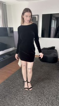 Μαύρο φόρεμα από βελούδο με μακριά μανίκια, εφαρμοστή γραμμή, μήκος μέχρι το μέσο του μηρού και λεία υφή. Φορεμένο με μαύρα πέδιλα με λουράκια, στέκεται πάνω σε ένα χαλί με υφή.