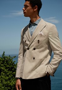 Beige double-breasted blazer with brown buttons, textured fabric, patch pockets, and a white pocket square over a light denim shirt.