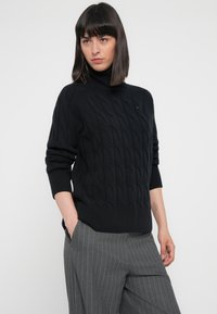 Maglione nero a maglia grossa con collo alto e orlo a coste. Il modello indossa pantaloni grigi a righe sottili, con le mani in tasca, con un branding minimale visibile.