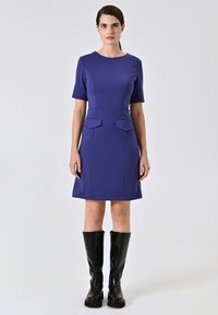 Paarse korte jurk met een ronde halslijn, zijzakken en een getailleerde taille, gecombineerd met kniehoogte zwarte laarzen. Gladde textuur.