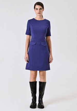 Lila kurzes Kleid mit Rundhalsausschnitt, seitlichen Taschen und taillierter Taille, kombiniert mit knielangen schwarzen Stiefeln. Glatte Textur.