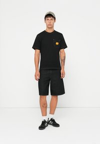 Μαύρο βαμβακερό t-shirt με μπροστινή τσέπη και πορτοκαλί ετικέτα, σε συνδυασμό με σκούρα τζιν σορτς. Μαύρα αθλητικά παπούτσια με λευκές ρίγες και λευκές κάλτσες.