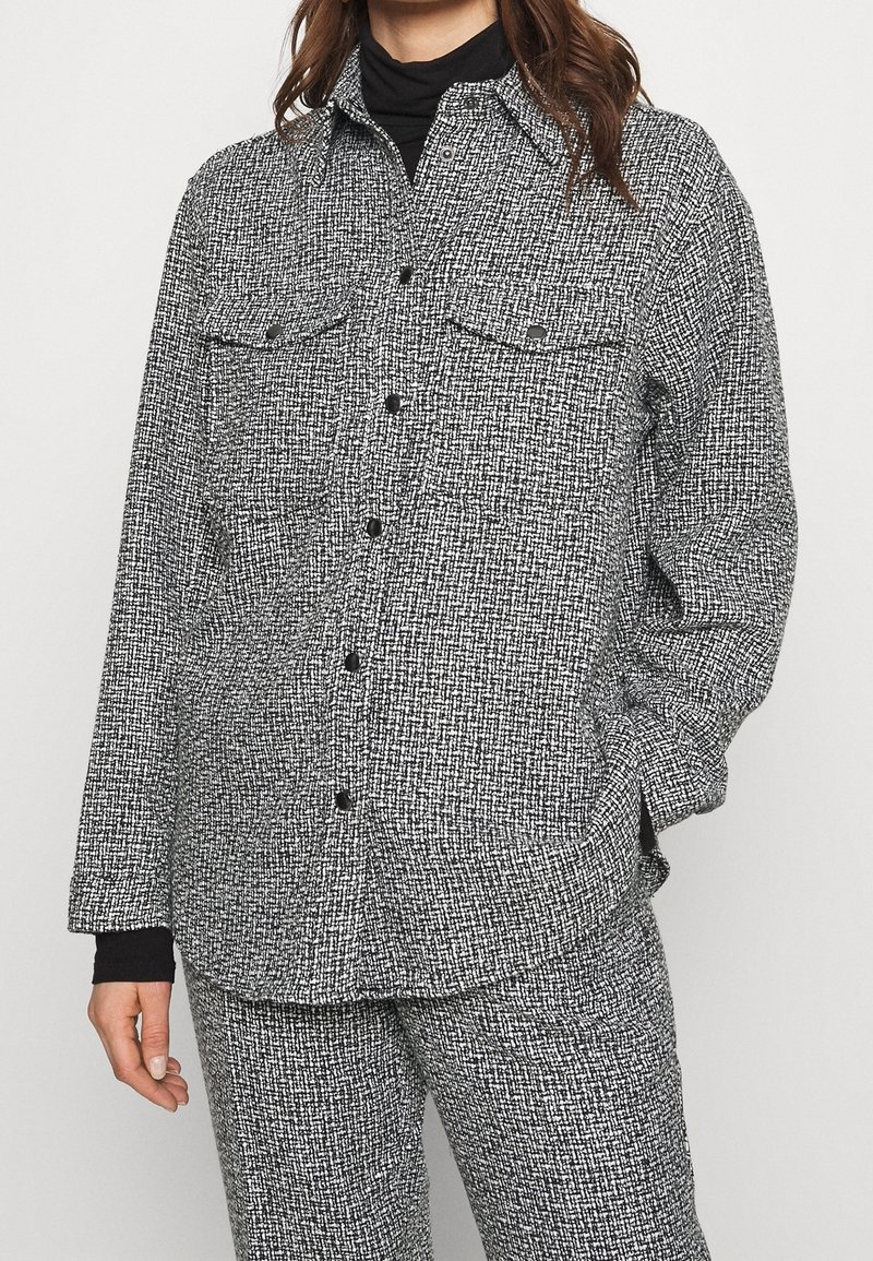 Femme portant une chemise boutonnée texturée noire et blanche assortie et un pantalon, avec un col roulé noir en dessous.